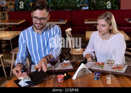 Man paying the bill with cash, after dinner with his girlfriend in a modern retro style restaurant. Fine dining and gentleman manners concept. Stock Photo