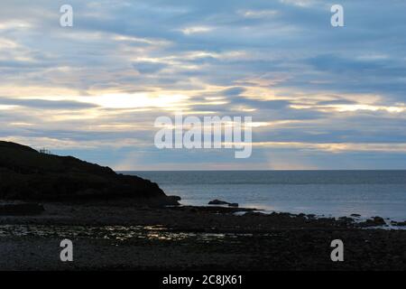 Sun rays coming through clouds onto the ocean, seen from a rocky beach in Trefor, North Wales Stock Photo