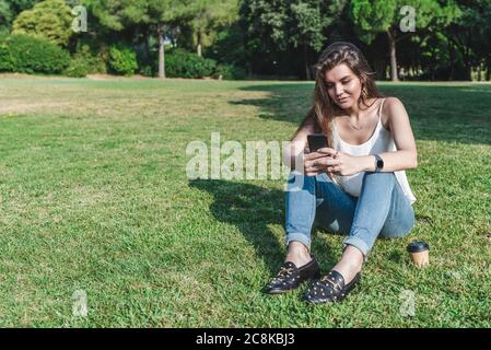 beautiful young caucasian woman sitting on the grass of an urban park texting with an smart phone Stock Photo
