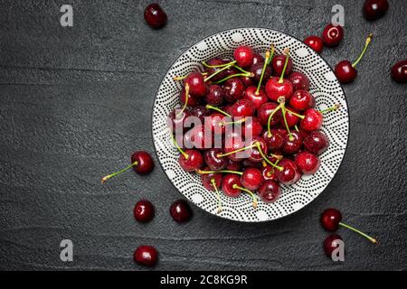 Juicy ripe cherries with water drops in ceramic bowl on black concrete table, top view with empty space.