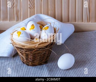 One white egg and other easter eggs in a small brown rough basket on a wooden background. Funny decorating with decoupage. Cute chickens. Stock Photo