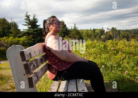 A woman sitting on a park bench beside grass blowing in the breeze on the east coast ocean Stock Photo