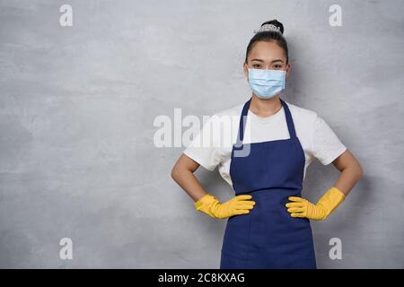 Cleaning services during pandemic. Portrait of a cleaning lady wearing yellow rubber gloves and medical protective face mask keeping arms on hips, looking at camera while standing against grey wall Stock Photo