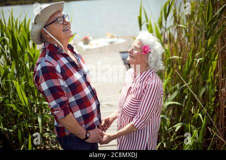 close up of cute caucasian senior couple holding hands, looking up, laughing,  standing by the river Stock Photo