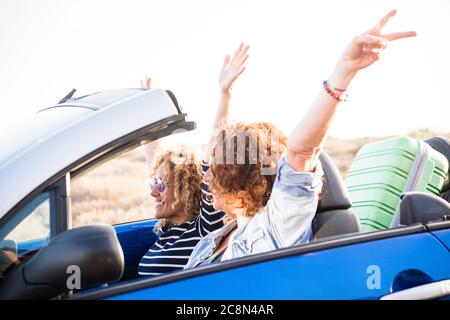 Happy travel adult friends lifestyle concept with couple of adult caucasian woman in a convertible car and luggage behind the seats - up arms and happ Stock Photo
