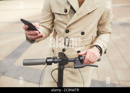 Cropped portrait of unrecognizable young man wearing trenchcoat holding smartphone while riding electric scooter in urban city setting, copy space Stock Photo