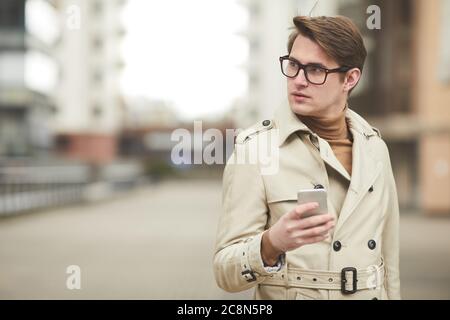 Minimal waist up portrait of modern young businessman wearing trenchcoat and holding smartphone looking away while posing confidently in urban city se Stock Photo