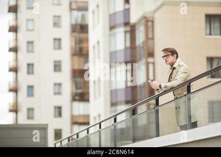 Graphic wide angle portrait of modern young businessman wearing trenchcoat and holding smartphone while leaning on railing outdoors in urban city sett Stock Photo