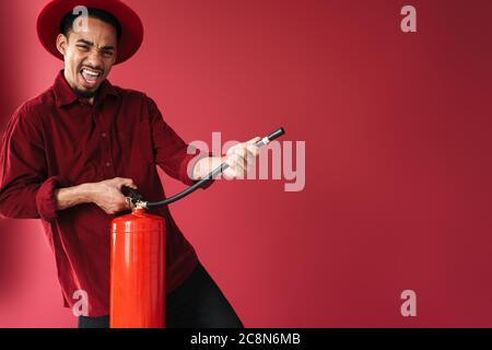 Image of a handsome african emotional young man posing isolated over red wall background holding fire extinguisher Stock Photo