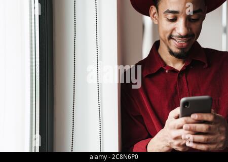 Handsome smiling stylish young african man using mobile phone while standing at the window indoors Stock Photo