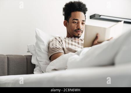 Photo of serious african american man with mustache reading book while lying in bed at bright room Stock Photo