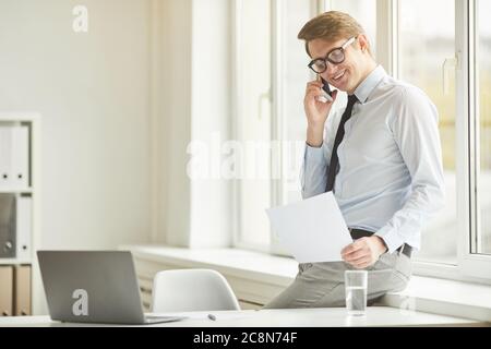 Side view portrait of cheerful young businessman speaking by smartphone while leaning on window at workplace in office, copy space Stock Photo