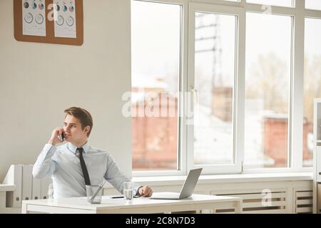 Minimal portrait of smiling young businessman speaking by smartphone and looking away while working at desk in white office interior, copy space Stock Photo