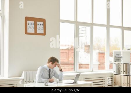 Wide angle portrait of exhausted young businessman rubbing forehead while suffering from migraine in white office interior, copy space Stock Photo