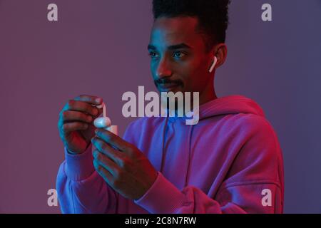 Portrait of casual african american man in colorful hoodie listening to music with earpod isolated over violet background Stock Photo