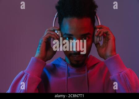 Portrait of casual african american man in colorful hoodie listening to music with headphones isolated over violet background Stock Photo