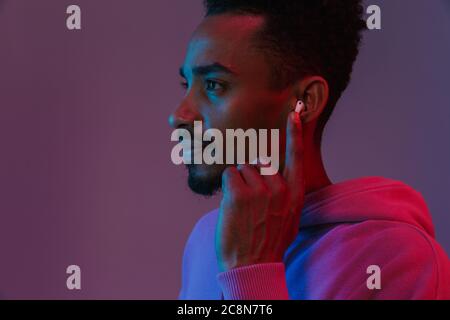 Portrait of concentrated african american man in colorful hoodie listening to music with earpod isolated over violet background Stock Photo