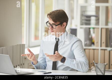 Portrait of young businessman looking at laptop screen and gesturing during video call in minimal office interior, copy space Stock Photo