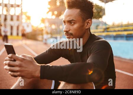 Image of young african american man holding smartphone while sitting at sports ground outdoors Stock Photo