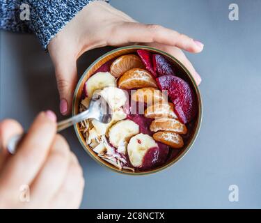 Acai smoothie, granola, seeds, fresh fruits in a wooden bowl in female hands on grey table. Eating healthy breakfast bowl. Top view Stock Photo