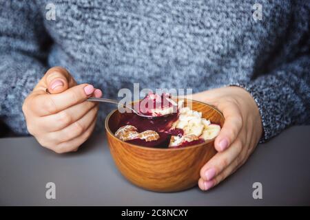 Acai smoothie, granola, seeds, fresh fruits in a wooden bowl in female hands on grey table. Eating healthy breakfast bowl Stock Photo