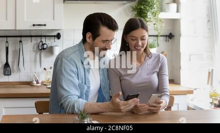 Smiling millennial couple relax on kitchen using smartphones Stock Photo