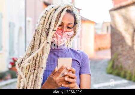 Yound african woman with blond dreadlocks using mobile phone while wearing face protective mask - Trendy person having fun with technology trends - Te Stock Photo