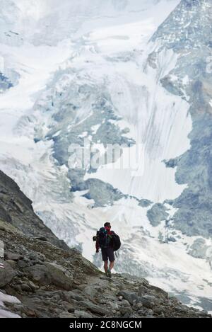Back view of alone tourist with backpack walking on rocks, beautiful mountains scenery on background. Trekking, man reaching peak. Wild nature with amazing views. Sport tourism in Alps. Stock Photo