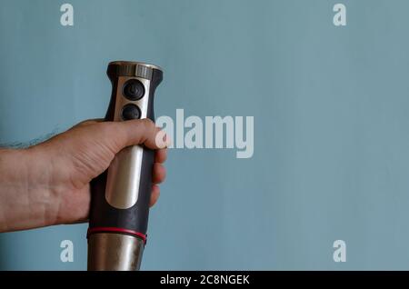 Hand holds a kitchen mixer with a nozzle. Left Hand of a man with an electric kitchen chopper. Caucasian man 43-45 years old. Kitchen appliances. Stock Photo