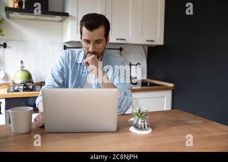 Pensive young man work on laptop in kitchen thinking Stock Photo