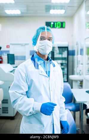 A medical worker, a laboratory assistant in a protective mask, visor, disposable cap and rubber gloves stands in a laboratory, hospital. Stock Photo