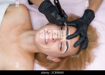 Cosmetology cabinet client lies on couch. Beautician applies permanent ink on eyebrows. Procedure of permanent eyebrow makeup. Close up black glove. Beauty industry Stock Photo