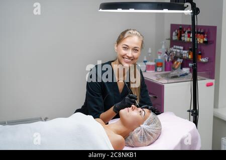 Cosmetology cabinet client lies on couch. Beautician applies permanent ink on eyebrows. Procedure of permanent eyebrow makeup. Free space. Beauty industry Stock Photo