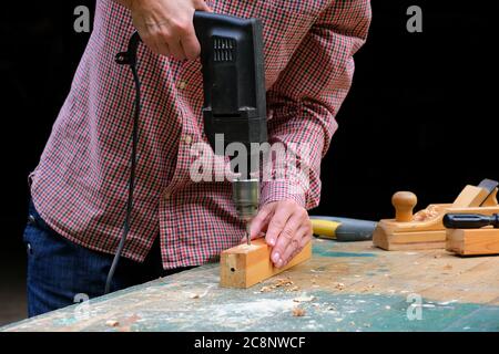 Craftswoman drilling a hole with an electric drill machine in wooden plank, closeup view. Concept of joiner's shop and woodworking. Gender equality. Stock Photo