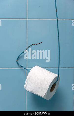 A roll of toilet paper hangs on a wire on a wall from blue tiles. Simple toilet paper holder at bathroom. Stock Photo
