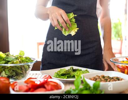 Woman chef preparing a fresh salad at home with sunlight in background with lettuce, tomatoes, beans, seeds on the table - Healthy vegan and vegetaria Stock Photo