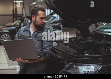 Experienced car technician using laptop, during car diagnostic at his workshop. Bearded male mechanic repairing modern SUV car at his garage, copy spa Stock Photo