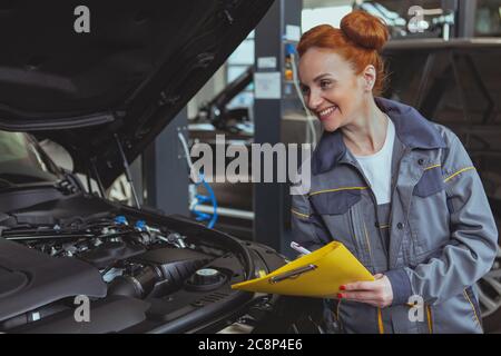 Beautiful woman working at car repair service, taking notes on clipboard. Attractive female technician examining engine of an auto. Female mechanic lo Stock Photo