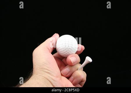Man's hand holding golf ball & golf tee on a black background. Stock Photo