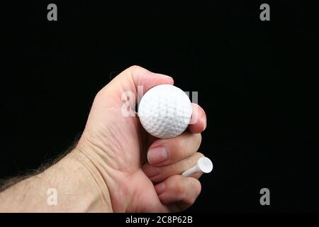 Man's hand holding golf ball & golf tee on a black background. Stock Photo