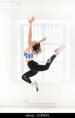 Woman doing a graceful acro gymnastic butterfly jump with outstretched arms in a high key gym in a health and fitness or active lifestyle concept Stock Photo