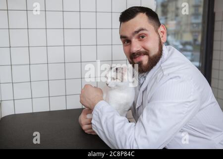 Cheerful male vet smiling to the camera, hugging cute white cat while working at his medical care office Stock Photo