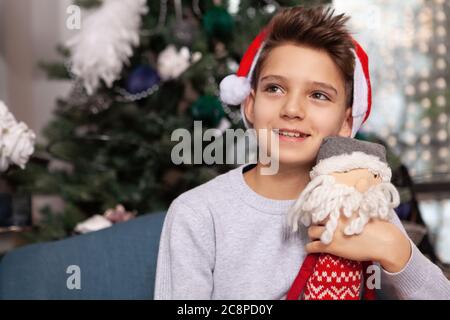 Charming young boy wearing Christmas hat, hugging his Santa Claus toy, smiling joyfully sitting near x-mas tree at home Stock Photo