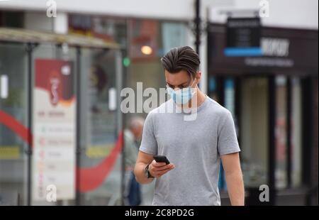 A young British man in his twenties wears a face mask in the street as he walks and texts on his smart phone Stock Photo