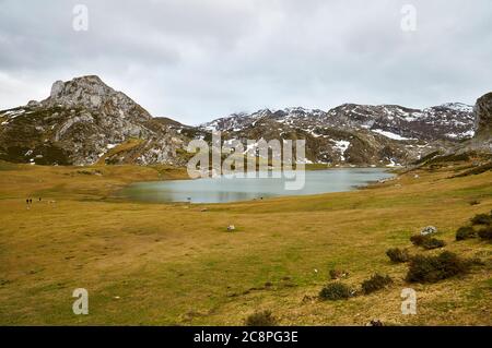 Hikers around Lago Ercina glacial lake in a cloudy day with snow in the nearby peaks (Cangas de Onís, Picos de Europa National Park, Asturias, Spain) Stock Photo