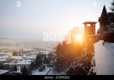 The bright setting sun shining between trees and buildings, overlooking a snow covered landscape Stock Photo
