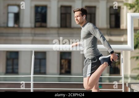 Young muscular man in sportswear and wireless headphones doing stretch for leg Stock Photo