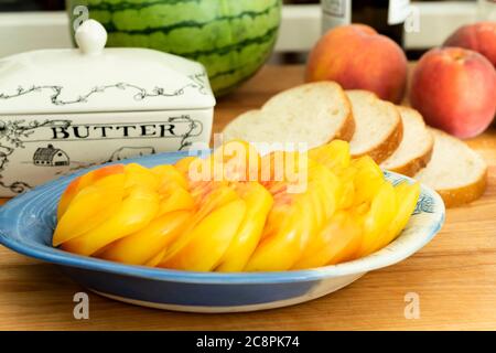 A yellow heirloom tomato, sliced, and arranged in a blue dish with sliced bread, a butter dish, and peaches. Stock Photo