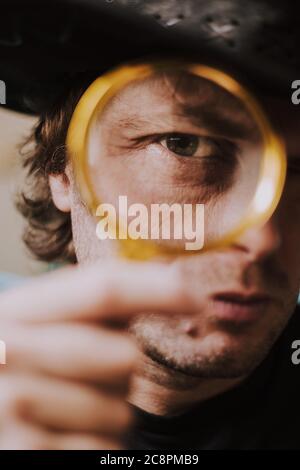 A discoverer with bristles on a brutal face and wearing a hat looks into the frame through a golden magnifying glass - naturalist and traveler Stock Photo