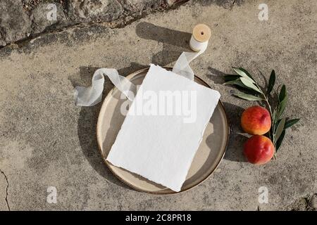 Summer wedding stationery. Blank greeting card mockup on ceramic plate in sunlight. Silk ribbon, peach fruit and olive branch on concrete background Stock Photo